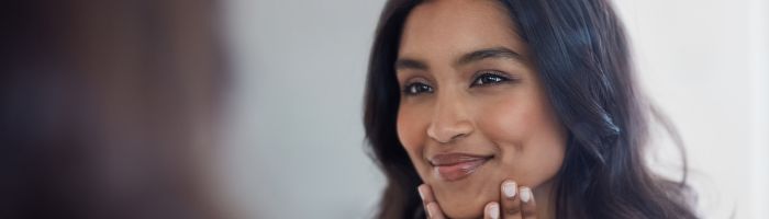 A woman smiling into a mirror, touching her chin with both hands.